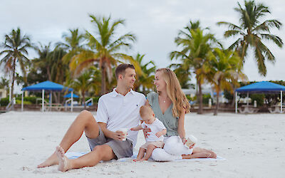 A family of three sits on a sandy beach with palm trees and blue canopies in the background, enjoying a relaxed moment together.