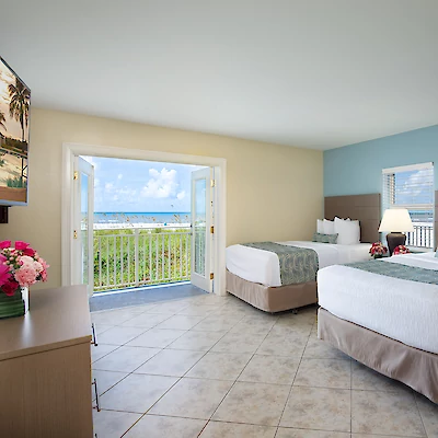 A hotel room with two beds, a TV, a dresser with flowers, and an open door with an ocean view and balcony railing outside.