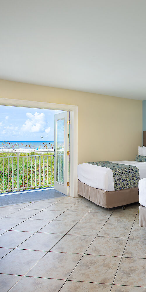 A hotel room with two beds, a TV, a dresser with flowers, and an open door with an ocean view and balcony railing outside.