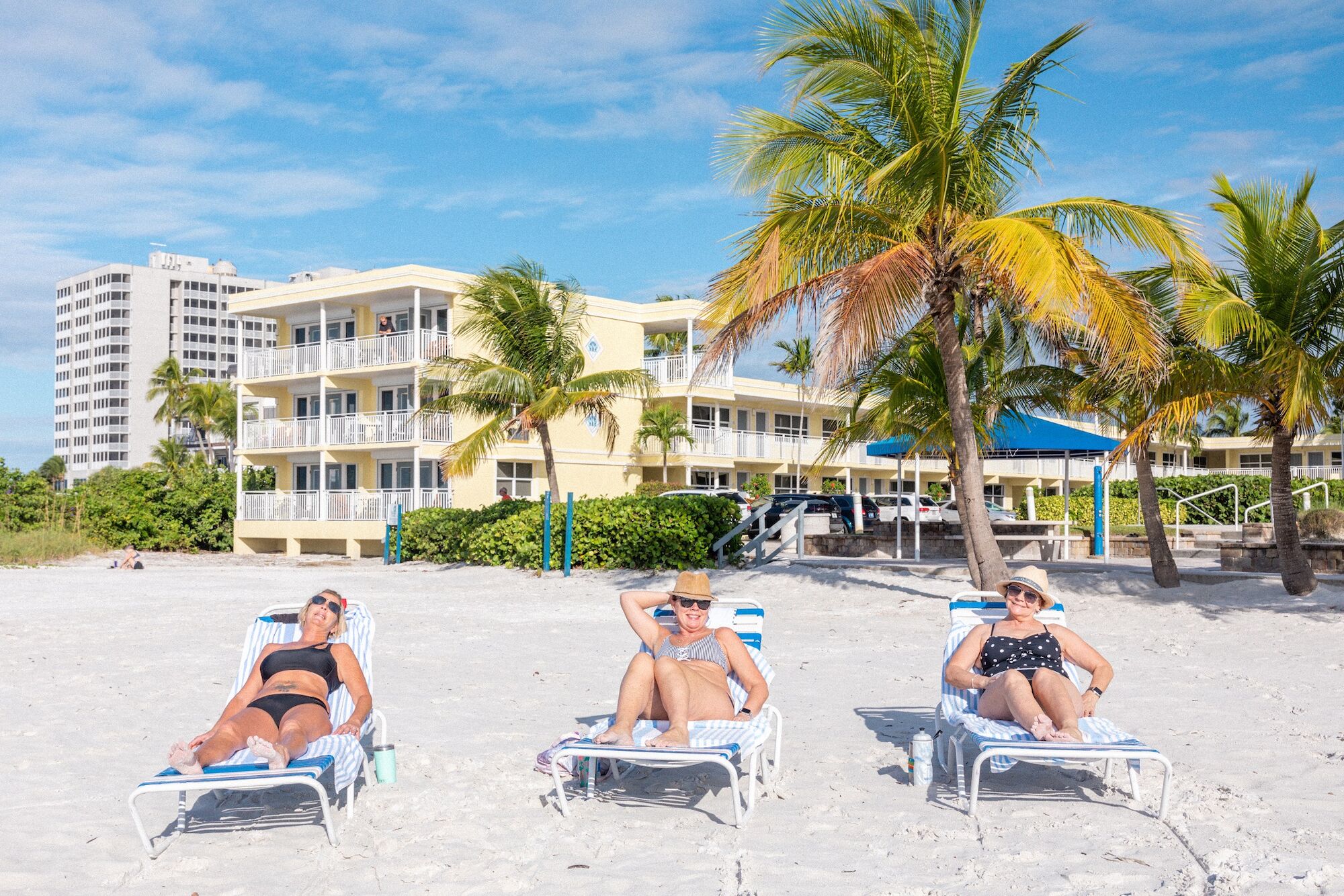 Three people are lounging on beach chairs under palm trees in front of a multi-story building under a clear blue sky.