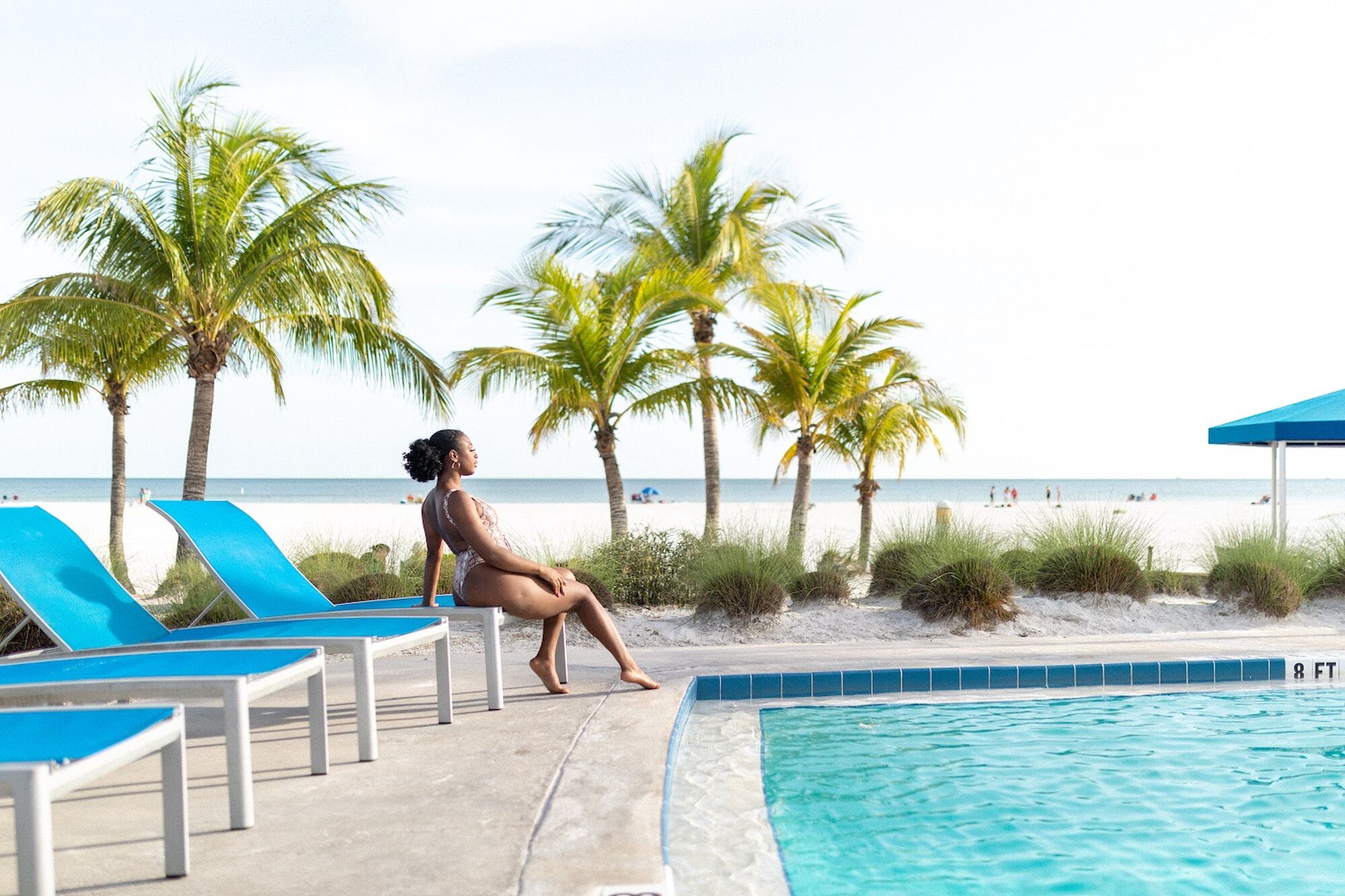 A person sits by a poolside with blue lounge chairs nearby, palm trees, and a beach in the background, looking at something in the distance.