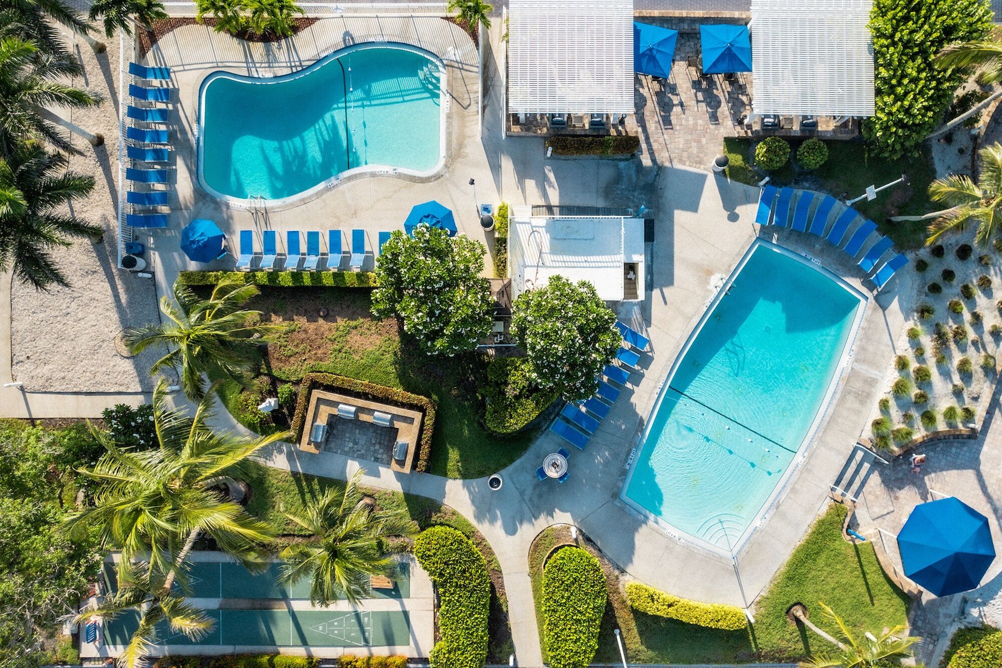 An aerial view of a resort pool area with two pools, lounge chairs, umbrellas, cabanas, palm trees, and greenery.