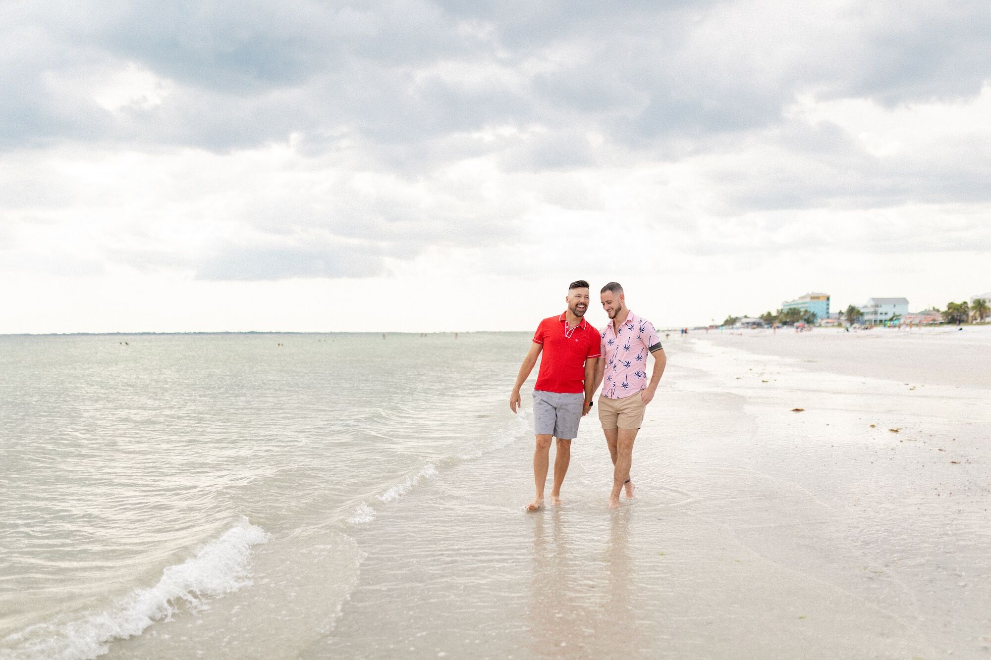 Two people are walking along a beach with calm waves under a cloudy sky. One wears a red shirt and shorts, the other a pink shirt and beige shorts.